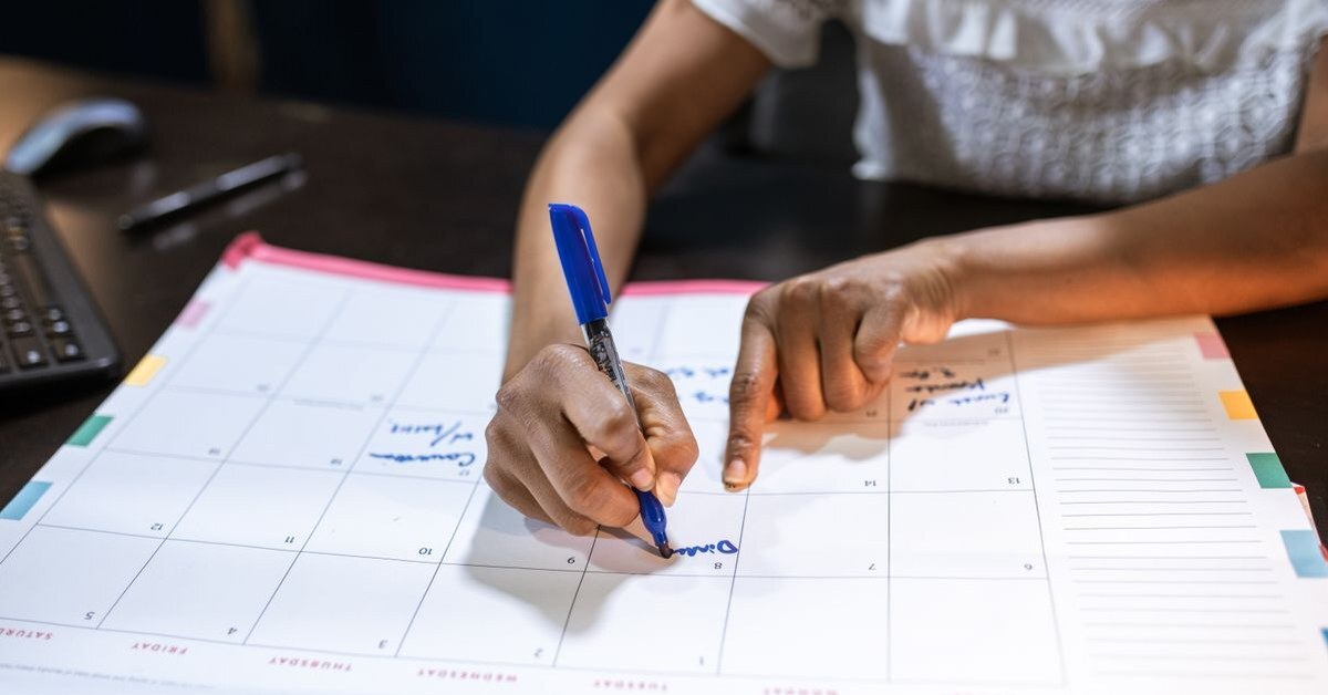A staff member writes a milestone in a blue marker on a large monthly desk planner, mapping out a step-by-step plan