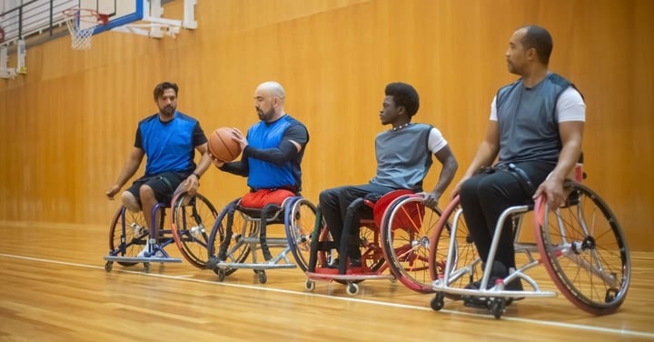 Four adults in sports wheelchairs on a gym court, one holding a basketball during an adaptive basketball session