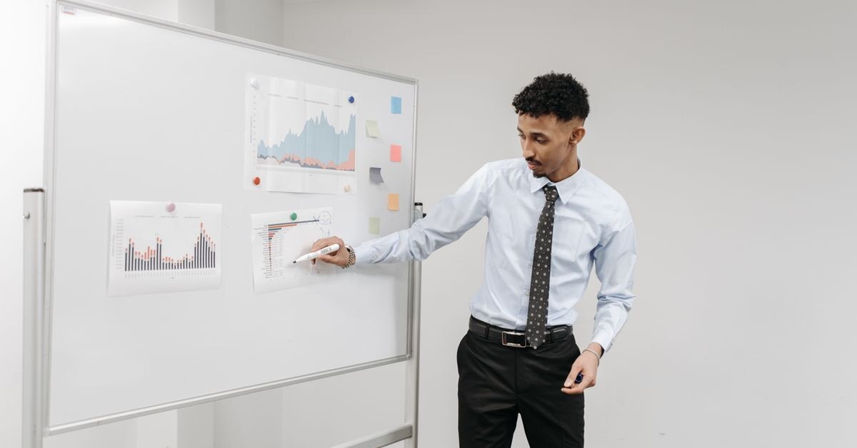 Staff member pointing at printed bar and area charts pinned to a whiteboard while presenting data to a room
