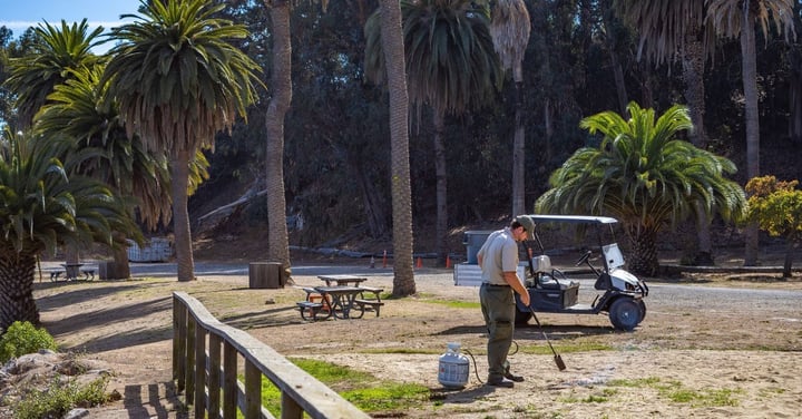 Park maintenance worker with a utility cart tending grounds by picnic tables, the routine upkeep that prevents costly repairs