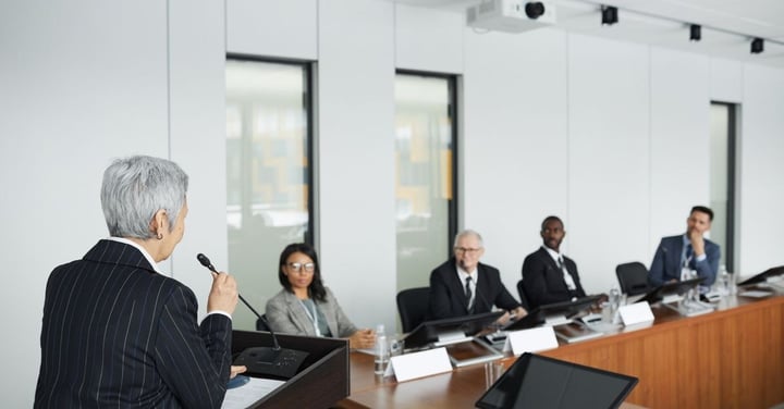 Woman speaking at a podium to a panel of officials seated at a long table in a meeting room
