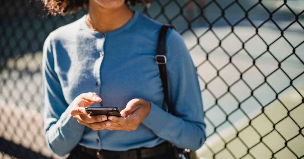 Woman holding a smartphone outdoors beside a chain-link fence, the mobile check-in families expect from a rec department