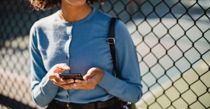 Woman holding a smartphone outdoors beside a chain-link fence, the mobile check-in families expect from a rec department