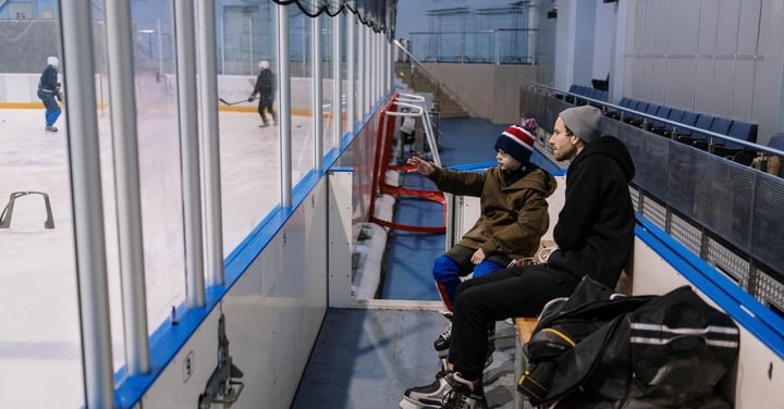 Father and son in winter hats sitting rinkside with a hockey bag while skaters practice behind the glass