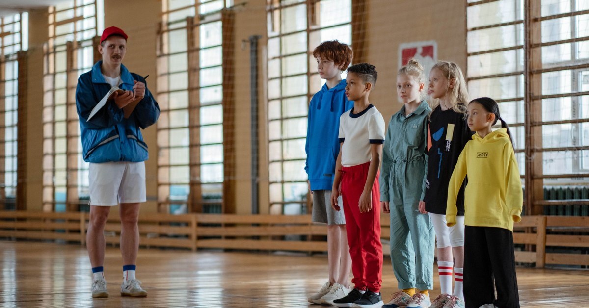 Youth sports leader taking notes in a notebook while a line of children waits in a school gym