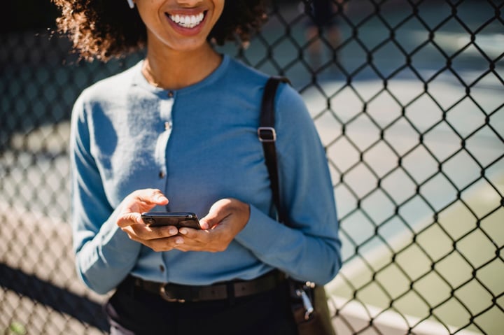 A cheerful woman uses her smartphone outdoors near a fence.